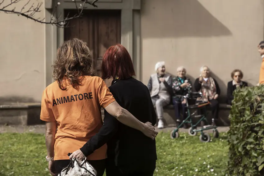 Anziani ospiti nel giardino della RSA Pio Istituto Santa Caterina di Prato, con un'animatrice vista di spalle che cammina abbracciata ad un'anziana